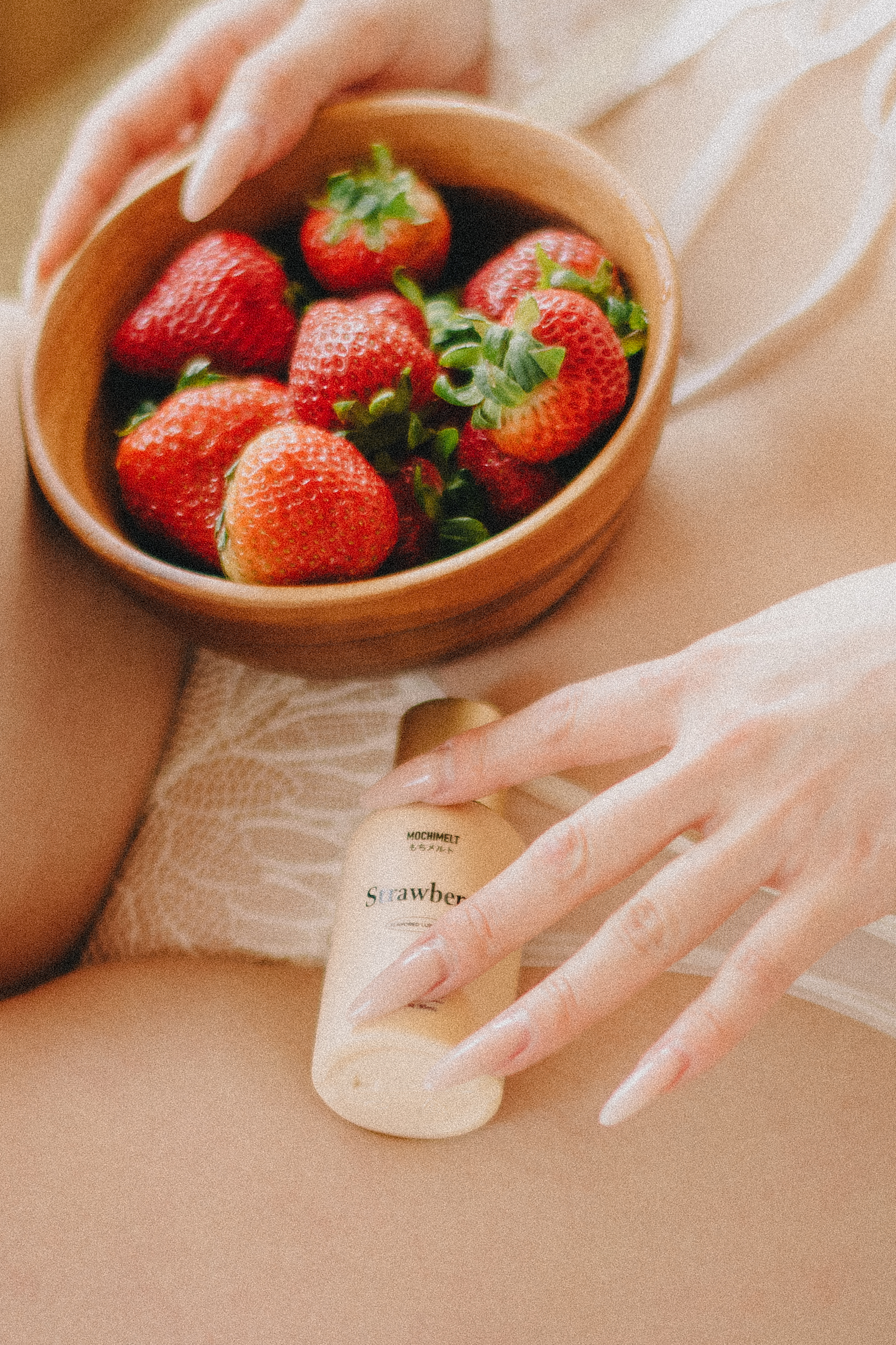 Mochi Melt strawberry lube next to a bowl of strawberries, on the lap of a woman wearing attractive lingerie