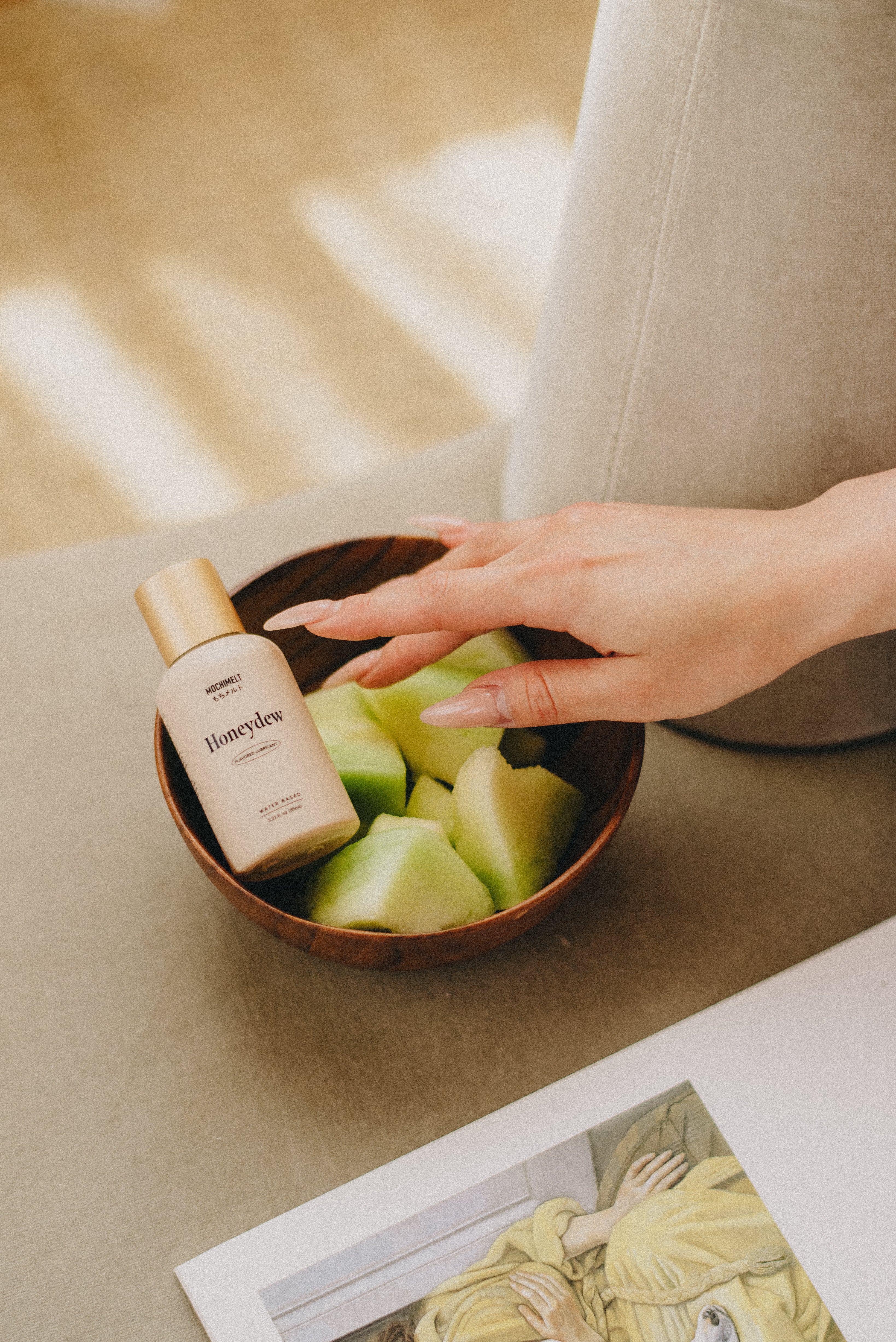Woman's well-manicured hand reaching for Honeydew fruit and Mochi Melt honeydew flavored lubricant in a wooden bowl